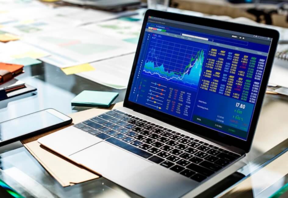 laptop on glass desk with papers showing trading chart